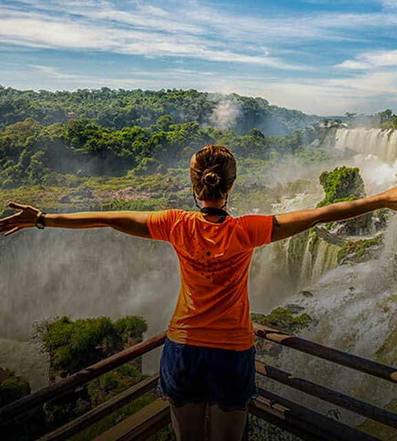 A woman in an orange shirt stands in front of Iguazu Falls and rainforest with outstretched arms.
